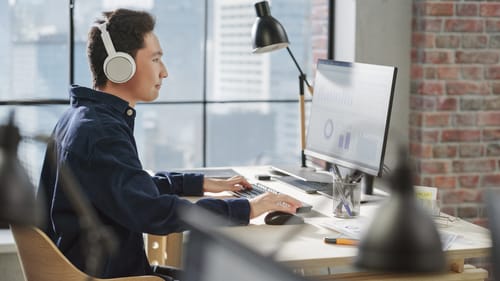 Man met hoofdtelefoon op werkt aan een bureau in een modern kantoor en kijkt naar grafieken op een computermonitor.