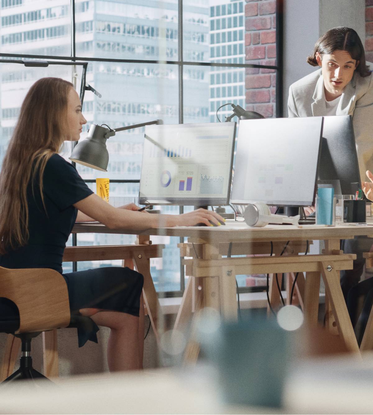 In een moderne kantoorruimte werken drie collega’s samen aan bureaus met computermonitoren met daarop grafieken; op de achtergrond grote ramen met stadsgezicht.