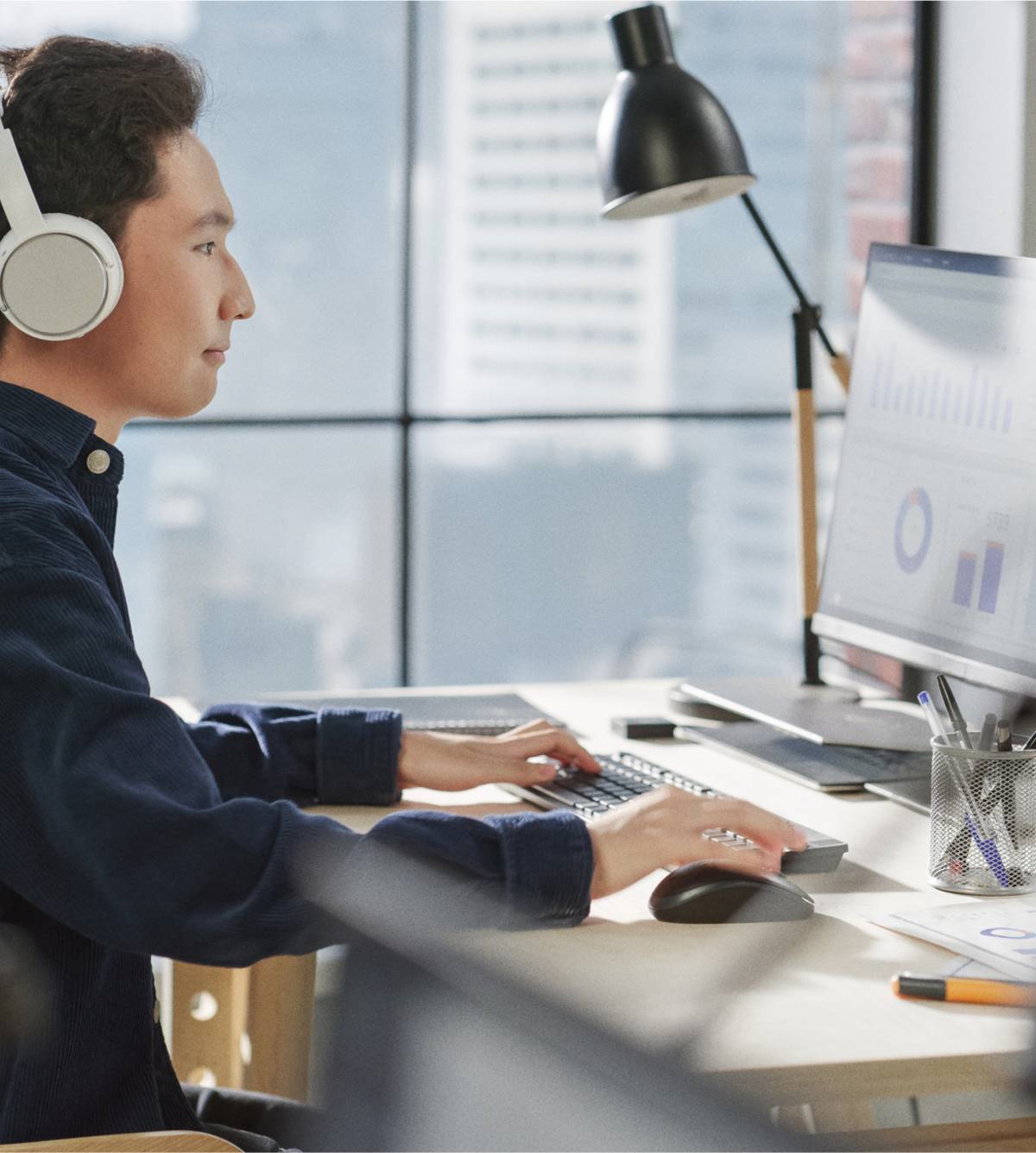 Man met hoofdtelefoon op werkt aan een bureau in een modern kantoor en kijkt naar grafieken op een computermonitor.